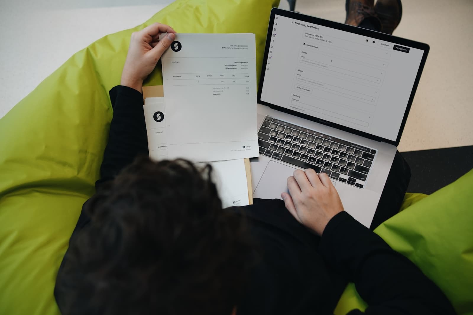 Someone working through printed invoices beside a laptop showing an invoicing screen