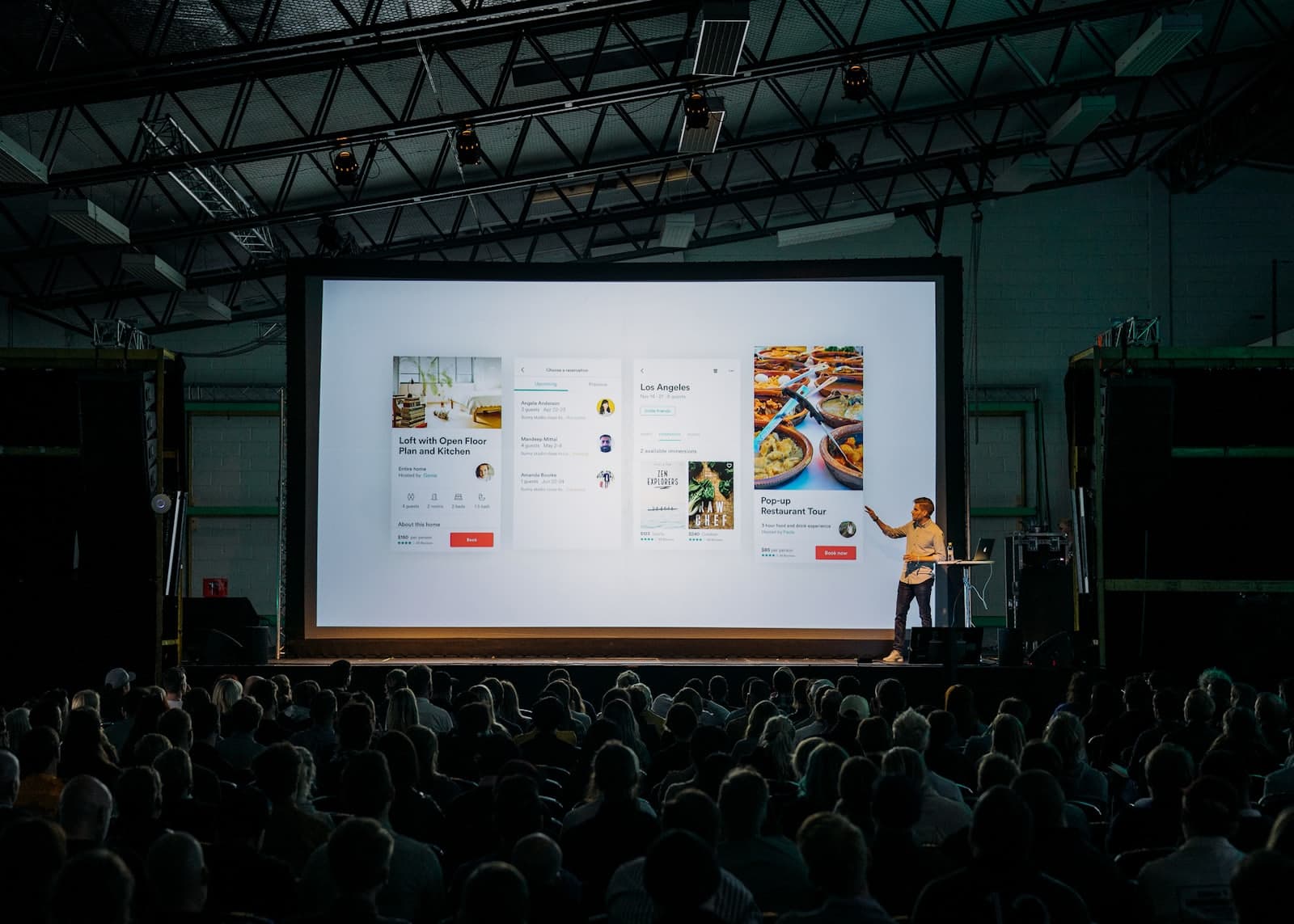 Person discussing while standing in front of a large screen in front of people inside dim-lighted room